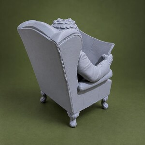 May include: A light gray 3D printed figurine of a figure seated in a high-backed wingback chair. The figure is holding an open book or newspaper. The chair features detailed upholstery textures, nailhead trim along the edges, and carved legs.