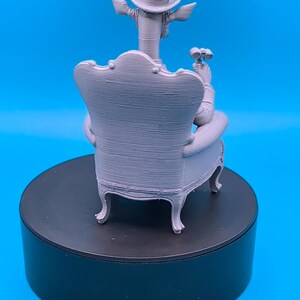 May include: A light gray 3D printed figurine of a giraffe wearing a top hat, seated in a high-backed armchair. The giraffe is holding a pair of binoculars in one hand. The figure displays visible horizontal layer lines from the printing process.