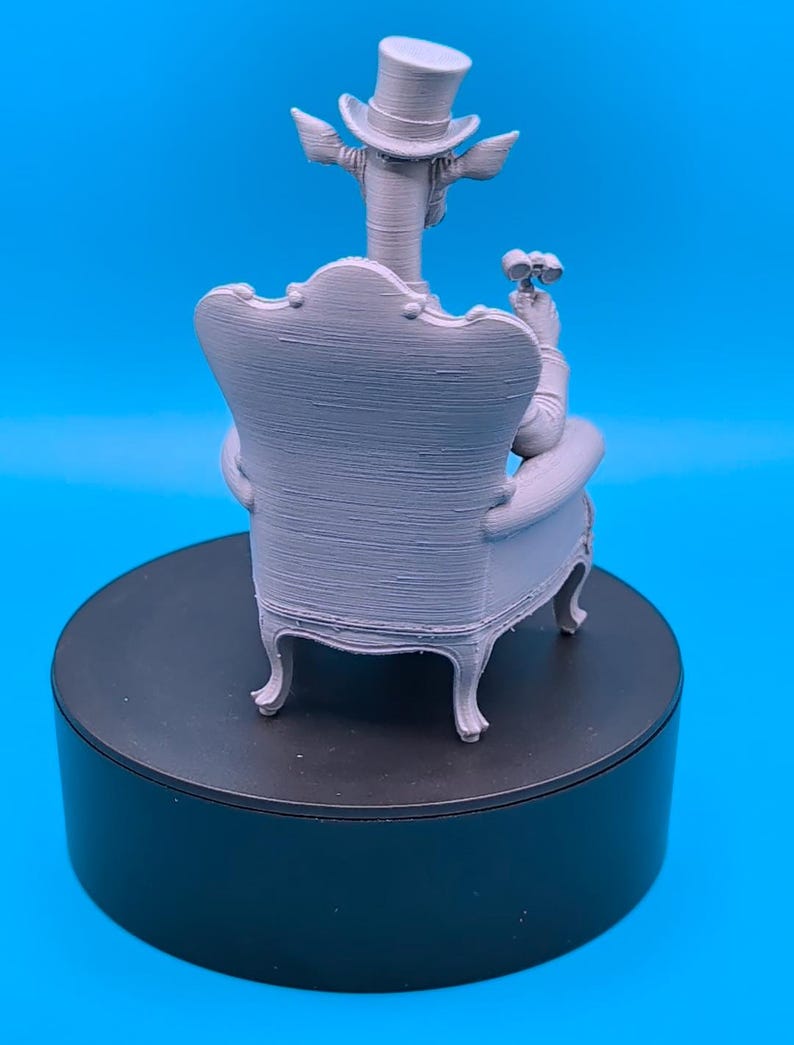 May include: A light gray 3D printed figurine of a giraffe wearing a top hat, seated in a high-backed armchair. The giraffe is holding a pair of binoculars in one hand. The figure displays visible horizontal layer lines from the printing process.