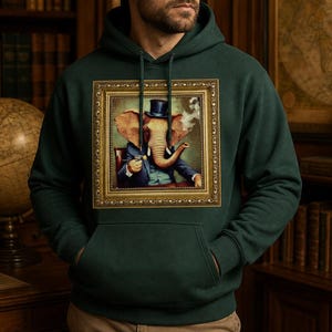 May include: A dark green hooded sweatshirt featuring a central graphic print of an elephant wearing a top hat and suit, depicted as if inside an ornate gold picture frame. The hoodie includes a front kangaroo pocket and matching green drawstrings.