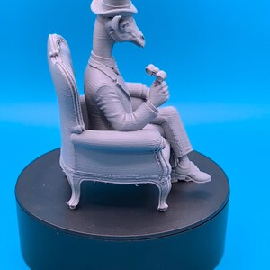May include: A light gray 3D printed figurine of an anthropomorphic giraffe wearing a formal suit and a top hat, seated in an armchair while holding a small object in one hand. The figure displays visible horizontal layer lines from the printing process.