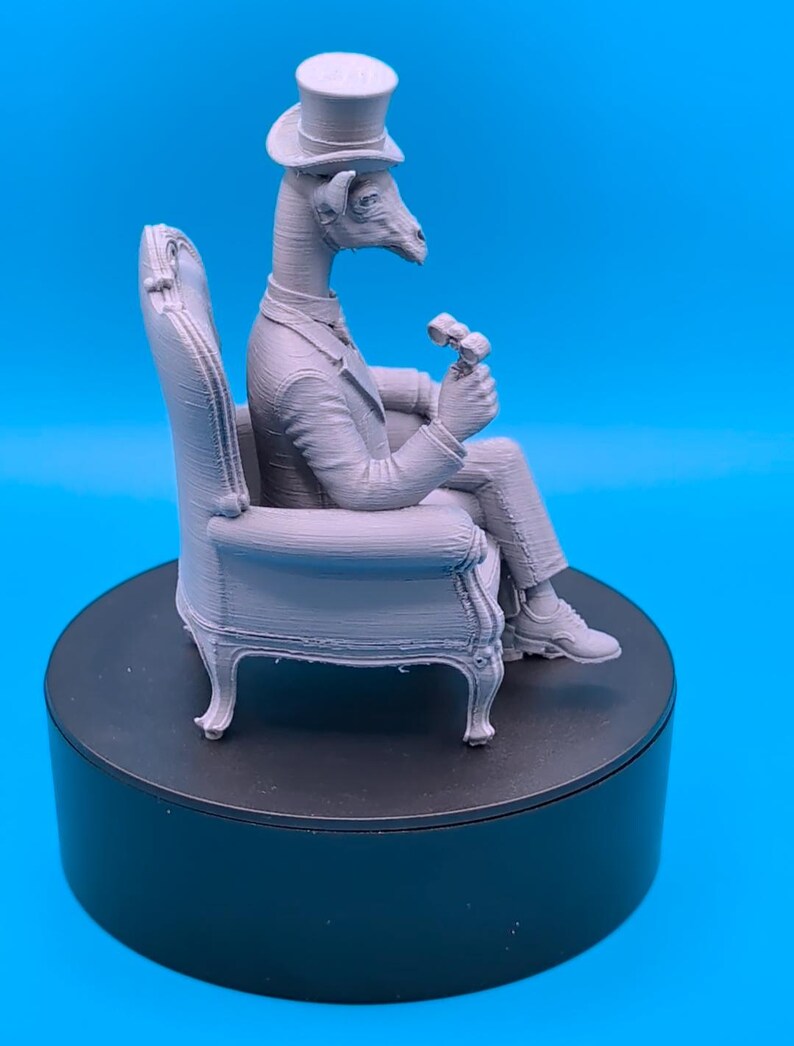 May include: A light gray 3D printed figurine of an anthropomorphic giraffe wearing a formal suit and a top hat, seated in an armchair while holding a small object in one hand. The figure displays visible horizontal layer lines from the printing process.
