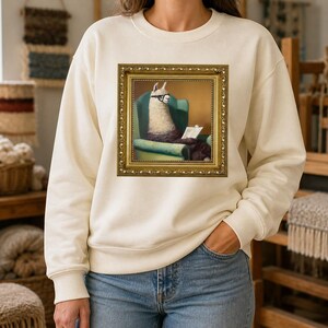 Puede incluir: Una sudadera de cuello redondo color crema con un estampado gráfico central de una llama con gafas, sentada en un sillón verde azulado y leyendo un libro, todo encerrado dentro de un marco rectangular de color dorado detallado.