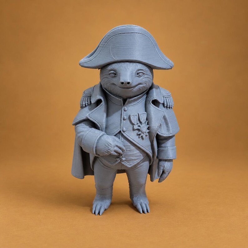 Puede incluir: Una figura gris impresa en 3D de un perezoso vestido como un general militar. El perezoso lleva un sombrero bicornio, un abrigo largo con charreteras y un chaleco con dos medallas prendidas en el pecho. La figura tiene un acabado mate y est&aacute; de pie.