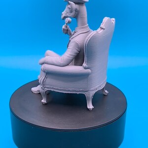 May include: A light gray 3D printed figurine of an anthropomorphic goat wearing a formal suit and a top hat, sitting in an ornate armchair while holding a small circular object. The figure is rendered in a monochromatic matte finish.