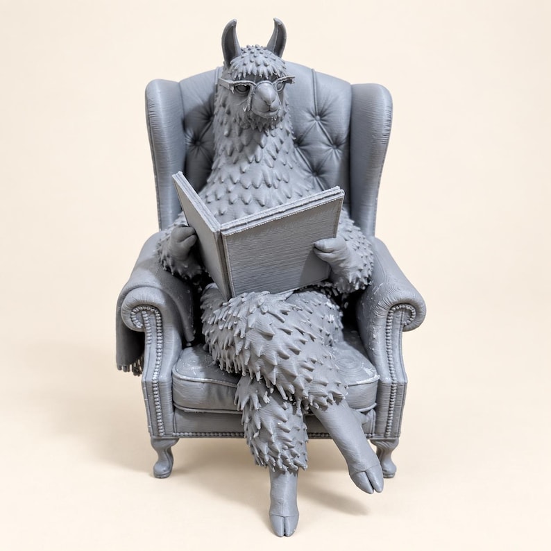K&ouml;nnte beinhalten: Eine einfarbige graue Lama-Figur mit Brille, die in einem gepolsterten Ohrensessel sitzt und ein offenes Buch h&auml;lt. Das Lama hat ein strukturiertes Fell und sitzt mit &uuml;berkreuzten Beinen in dem Sessel, der mit Knopfdetails verziert ist.
