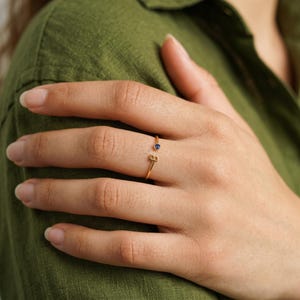 Op de afbeelding: Een goudkleurige open ring met een kleine, ronde, donkerblauwe edelsteen aan het ene uiteinde en de hoofdletter B aan het andere uiteinde. De ringband is dun en metaalachtig en ontworpen om de vinger gedeeltelijk te omsluiten met een opening tussen de letter en de steen.