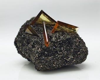 Op de afbeelding: Een donkere grijze rotsformatie met verschillende amberkleurige, geometrische kristalstructuren die uit het oppervlak steken. De kristallen hebben een doorschijnend uiterlijk en de rots heeft een ruw, getextureerd oppervlak.