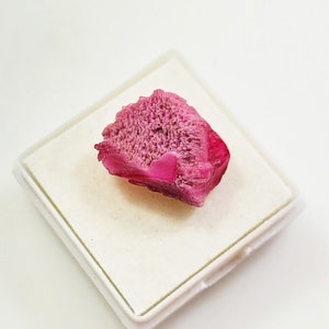 Könnte beinhalten: Ein leuchtend pinkfarbenes, kristallines Mineralstück mit einer rauen, strukturierten Oberfläche. Das Mineral wird auf einer weißen quadratischen Präsentationsbox gezeigt.