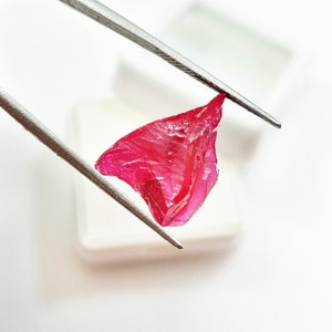 Könnte beinhalten: Ein leuchtend rosa Edelstein mit einer rauen, unregelmäßigen Form, gehalten von einer Pinzette.