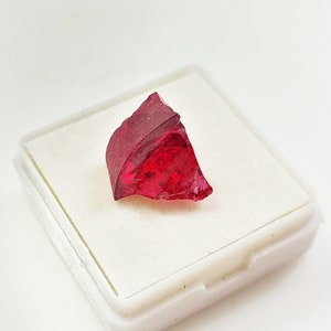 May include: A deep red, faceted gemstone in a white display box. The gemstone has a rough, natural shape.