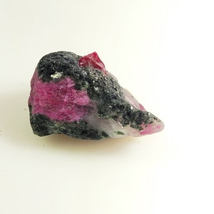 Peut inclure: Une pierre précieuse brute avec un cristal de rubis rouge vif incrusté dans une matrice sombre et mouchetée. La pierre présente un mélange de teintes rose foncé et noires, avec une structure cristalline.