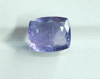 May include: A faceted, cushion-cut gemstone with a deep violet-blue hue. The gem displays multiple facets that reflect light, creating a sparkling effect. The stone is set against a light-colored background.