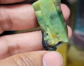 May include: A raw, uncut green gemstone with a crystalline structure. The gem has a translucent quality with areas of darker green and hints of orange. The base of the stone is a dark, almost black color. The gemstone is held in a hand.