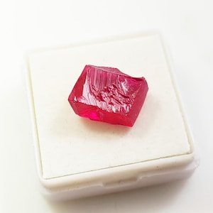 Puede incluir: Una piedra preciosa de rubí rojo intenso con un corte facetado, expuesta en una caja cuadrada blanca.