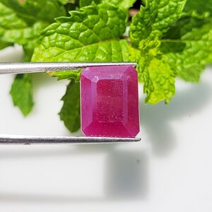 May include: A vibrant pink emerald-cut gemstone, held by a pair of tweezers, against a backdrop of green leaves.