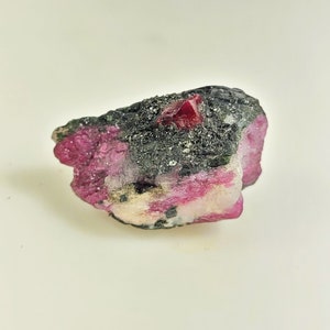 May include: A raw ruby gemstone with a deep red crystal structure. The ruby is set in a matrix of dark grey and black rock, with pink and white mineral deposits visible. The rough stone has a natural, unpolished appearance.
