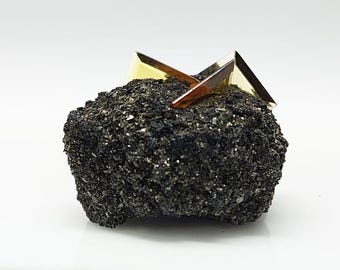 Puede incluir: Una formación rocosa oscura y texturizada con cristales metálicos de color dorado. Los cristales tienen forma geométrica, con bordes afilados y superficies reflectantes. La roca tiene un aspecto rugoso y granulado.