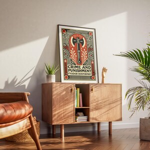 Op de afbeelding: Een ingelijste poster met een zwart-witte illustratie van een bijl op een rode en gouden achtergrond. De poster luidt "Misdaad en straf" van Fjodor Dostojevski.