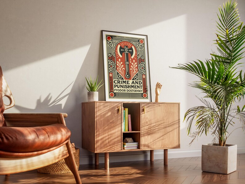 Op de afbeelding: Een ingelijste poster met een zwart-witte illustratie van een bijl op een rode en gouden achtergrond. De poster luidt "Misdaad en straf" van Fjodor Dostojevski.