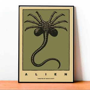 Puede incluir: Un póster enmarcado con un borde negro, que muestra una ilustración en negro y verde oliva de un facehugger alienígena. La palabra "ALIEN" está impresa en mayúsculas en la parte inferior, con "DIRECTED BY RIDLEY SCOTT" debajo.