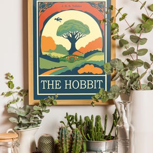 K&ouml;nnte beinhalten: Ein gerahmtes Poster mit einer Vintage-Illustration eines Baumes und eines Hobbit-Lochs. Das Poster tr&auml;gt den Titel "The Hobbit" und zeigt den Namen des Autors, J.R.R. Tolkien. Das Poster h&auml;ngt an einer wei&szlig;en Wand mit Pflanzen und einer Kamera im Vordergrund.