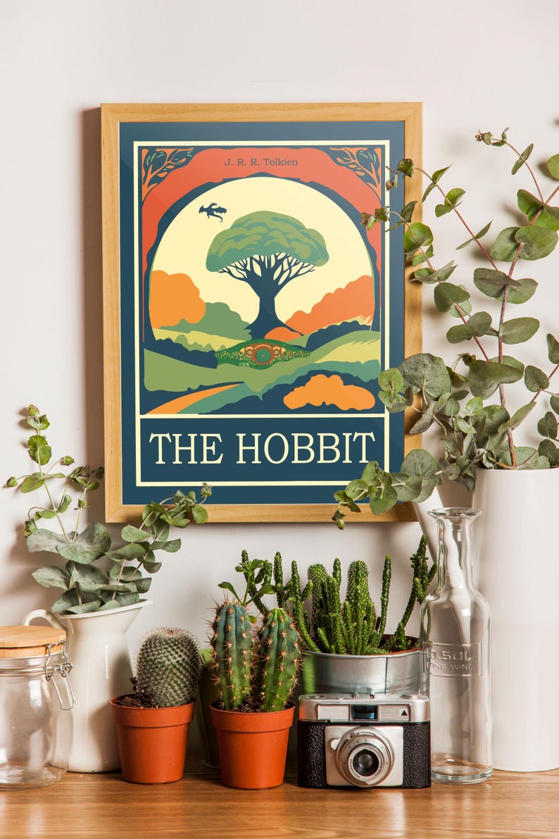 K&ouml;nnte beinhalten: Ein gerahmtes Poster mit einer Vintage-Illustration eines Baumes und eines Hobbit-Lochs. Das Poster tr&auml;gt den Titel "The Hobbit" und zeigt den Namen des Autors, J.R.R. Tolkien. Das Poster h&auml;ngt an einer wei&szlig;en Wand mit Pflanzen und einer Kamera im Vordergrund.