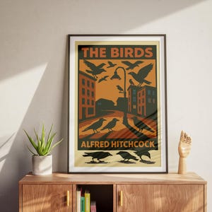 May include: A vintage-style poster with a dark brown frame featuring a black and white illustration of birds against a yellow and orange background. The poster reads "The Birds" and "Alfred Hitchcock".