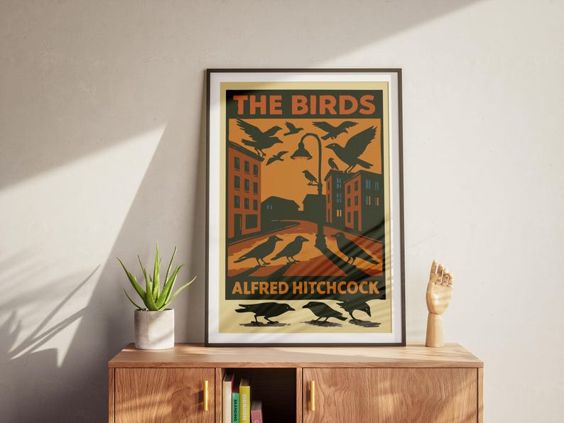 May include: A vintage-style poster with a dark brown frame featuring a black and white illustration of birds against a yellow and orange background. The poster reads "The Birds" and "Alfred Hitchcock".