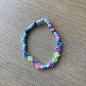 Könnte beinhalten: Ein kreisförmiges Armband aus kleinen, bunten Kunststoffperlen in Blau-, Lila-, Rosa- und Grüntönen. Mehrere Perlen sind mit Buchstaben in einer Kontrastfarbe bedruckt, die zusammen den Schriftzug WHAT COLOR ergeben.