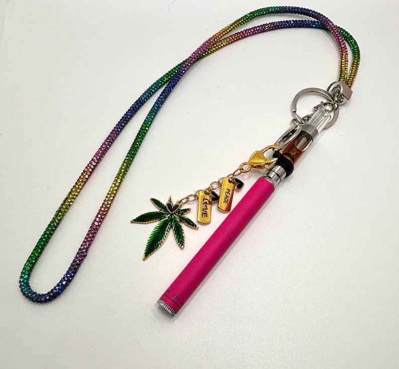May include: A pink vape pen with a clear glass tank and a rainbow lanyard. The lanyard has charms that say "LOVE", "PEACE", and a green marijuana leaf.
