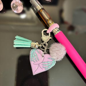 May include: A pink heart-shaped keychain with a skeleton hand design. The keychain is attached to a pink vape pen with a clear glass cartridge. The keychain also has a small, light blue tassel.