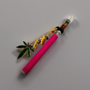 May include: A pink vape pen with a clear glass mouthpiece and a gold chain with charms that say "Peace", "Love", and a green marijuana leaf.