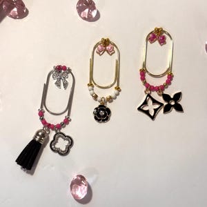 May include: Three decorative paper clips with various charms. One clip has a black tassel and a silver charm. Another has a black flower charm, and the third has black and white charms. All have pink and gold accents.