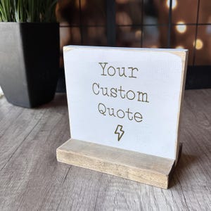 May include: A small, rectangular wooden sign with a white painted surface and a natural wood base. The sign displays the text "Your Custom Quote" in brown lettering, with a lightning bolt symbol below. The sign is designed to stand upright.