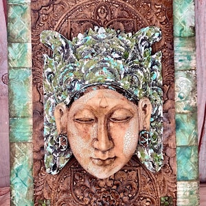 Può includere: Un'opera d'arte murale dettagliata e strutturata con un volto sereno con gli occhi chiusi, adornato da un copricapo decorativo. L'opera è principalmente marrone, con accenti di verde e turchese, ed è montata su una cornice di legno.