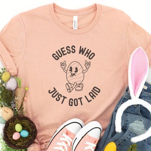 以下が含まれることがあります： 桃色のTシャツに「誰が産まれたか当ててごらん」という文字と、腕と脚のある漫画の卵が描かれています。卵は靴を履いていて、笑顔をしています。Tシャツは、卵の入った巣、ウサギの耳、青いピープなど、イースターをテーマにした装飾で囲まれています。