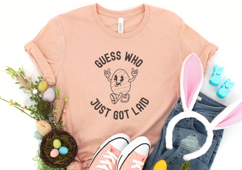 以下が含まれることがあります： 桃色のTシャツに「誰が産まれたか当ててごらん」という文字と、腕と脚のある漫画の卵が描かれています。卵は靴を履いていて、笑顔をしています。Tシャツは、卵の入った巣、ウサギの耳、青いピープなど、イースターをテーマにした装飾で囲まれています。