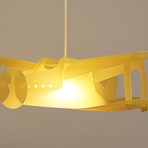 May include: A yellow airplane-shaped ceiling light fitting with a light bulb illuminating the inside. The airplane has a propeller and a small window.