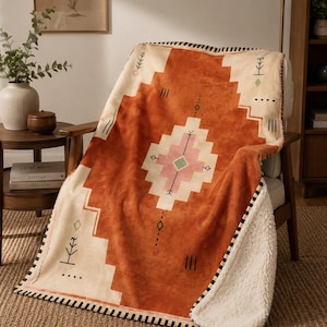 May include: A soft throw blanket featuring a rust-orange and cream geometric pattern with small pink and green accents. The blanket has a textured white sherpa fleece lining and a black and white striped edge trim along the perimeter.