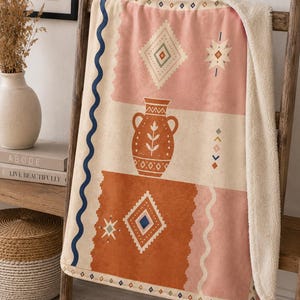 May include: A plush throw blanket featuring a cream-colored sherpa lining and a printed top side with a terracotta, pink, and cream color palette. The design includes a central terracotta vase, geometric diamond shapes, and wavy blue lines.