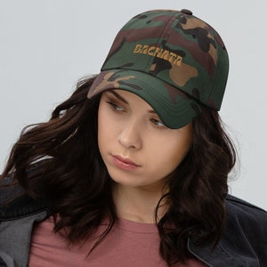 Puede incluir: Una gorra de béisbol de camuflaje con la palabra "BACHATA" bordada en dorado en la parte delantera.