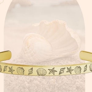 May include: Gold bangle bracelet with a delicate design of seashells and starfish. The bracelet is on a background of sand and a seashell.
