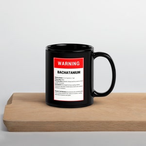 Puede incluir: Taza de café negra con una etiqueta de advertencia roja que dice "WARNING BACHATANIUM" y una descripción del estilo de baile. La taza está sobre una superficie de madera.