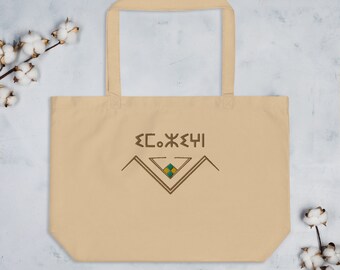 May include: A beige tote bag with a brown graphic design featuring a stylized mountain range and a diamond shape with yellow and green accents. The text "ΣC.ЖΞЧI" is printed above the graphic.