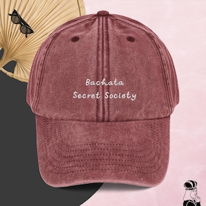 Könnte beinhalten: Eine weinrote Baseballkappe im Vintage-Stil mit weiß gesticktem Text, der "Bachata Secret Society" lautet.