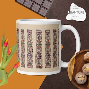 May include: White ceramic mug with a beige background and a repeating pattern of vertical diamond shapes in red, orange, green, and purple. The mug has a white handle and the word "QUANTURE" is visible on the side.