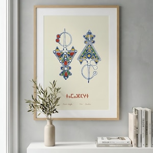 May include: Framed print featuring two ornate, symmetrical brooch designs in red, blue, yellow, and green. The artwork includes text in a stylized font and is displayed on a cream-colored background. The print is in a light wood frame.