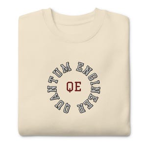 Könnte beinhalten: Ein cremefarbenes T-Shirt mit einem grauen gestickten Kreis, auf dem "Quantum Engineer" steht, wobei die Buchstaben "QE" in Rot im Kreis sind.