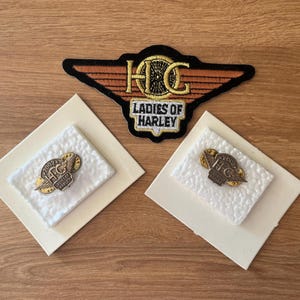 Puede incluir: Un surtido de parches y pines de Harley-Davidson Ladies of Harley. El parche es negro, naranja y dorado, con el texto "Ladies of Harley". También se ven dos pines, cada uno con el logotipo "H-D" y el texto "Ladies of Harley."